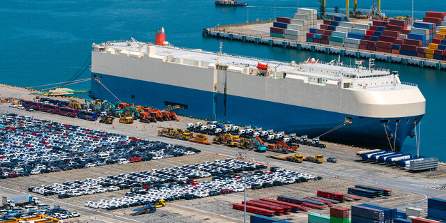 Tausende Autos auf einem Parkplatz an einem Hafen, ein Transportschiff liegt vor Anker.