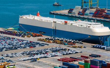 Tausende Autos auf einem Parkplatz an einem Hafen, ein Transportschiff liegt vor Anker.