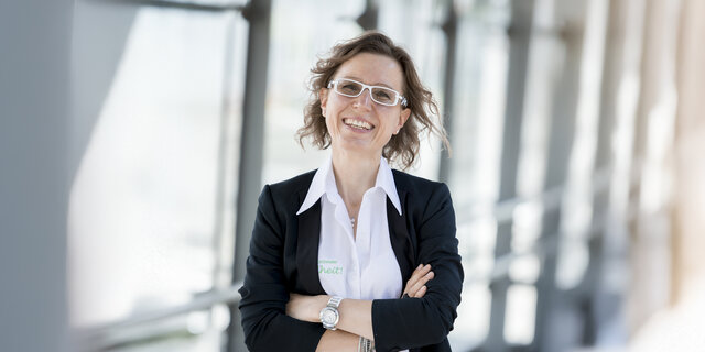 Ernährungsexpertin Angelika Kirchmaier steht vor Glasfenstern mit verschränkten Händen und lacht.