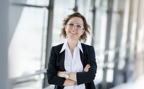 Ernährungsexpertin Angelika Kirchmaier steht vor Glasfenstern mit verschränkten Händen und lacht.