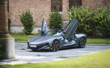 Ein graues Cabrio steht mit offenem Verdeck und Flügeltüren vor einem Backstein-Gebäude.