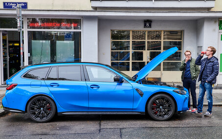 Ein blauer BMW M3 steht mit geöffneter Motorhaube auf einem Parkplatz, zwei Personen stehen neben dem Auto.