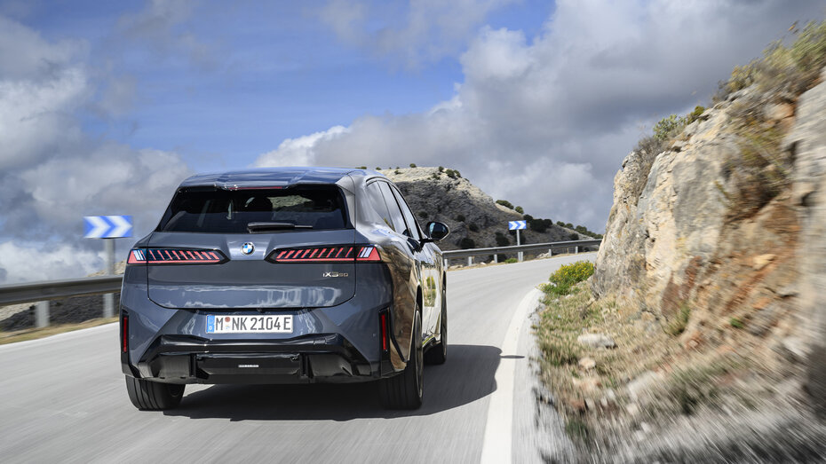 Ein BMW dunkelgrauer BMW iX3 fährt auf einer Landstraße an einer felsige Landschaft vorbei.