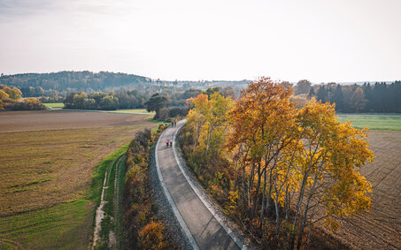 Bahntrassen-Radweg aus der Vogelperspektive. Es ist eine herbstliche Wettersituation, man sieht klein ein paar Fahrradfahrer:innen.