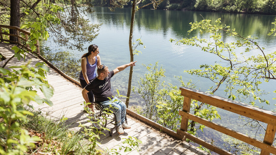 Eine Frau schiebt einen Mann im Rollstuhl auf einem barrierefreien Wanderweg am Reintalersee entlang.