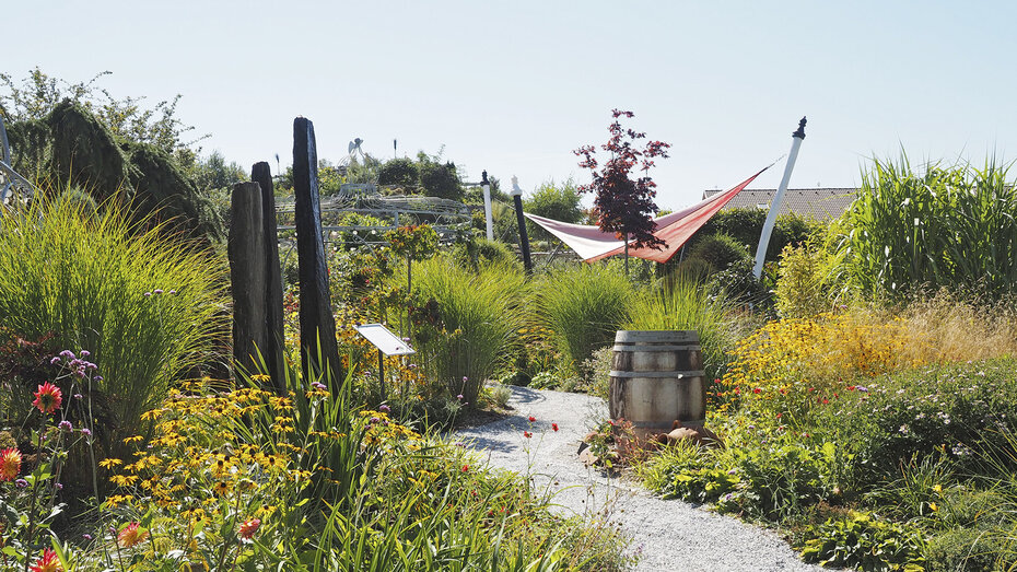 Brunnengarten in den Kittenberger Erlebnisgärten: Ein Weg führt durch grüne Blumen und Gräser zu einem mit Sonnensegel überdachtem Platz.