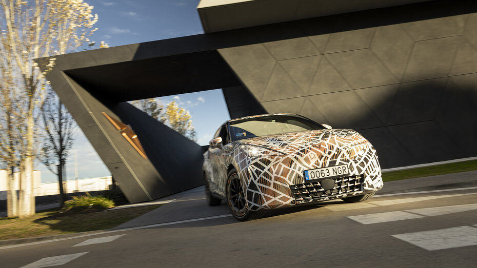 Ein mit Tarnfolie folierter Cupra Raval fährt bei einem futuristischen Gebäude vorbei.