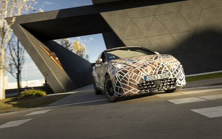 Ein mit Tarnfolie folierter Cupra Raval fährt bei einem futuristischen Gebäude vorbei.