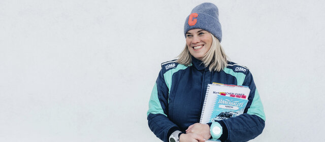 Catharina Schmidt, Rallye-Co-Pilotin und ÖAMTC-Mitarbeitetin steht im Renndress vor hellgrauem Hintergrund und hält ihre Rennnotizen in der Hand.