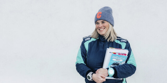 Catharina Schmidt, Rallye-Co-Pilotin und ÖAMTC-Mitarbeitetin steht im Renndress vor hellgrauem Hintergrund und hält ihre Rennnotizen in der Hand.