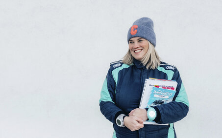 Catharina Schmidt, Rallye-Co-Pilotin und ÖAMTC-Mitarbeitetin steht im Renndress vor hellgrauem Hintergrund und hält ihre Rennnotizen in der Hand.