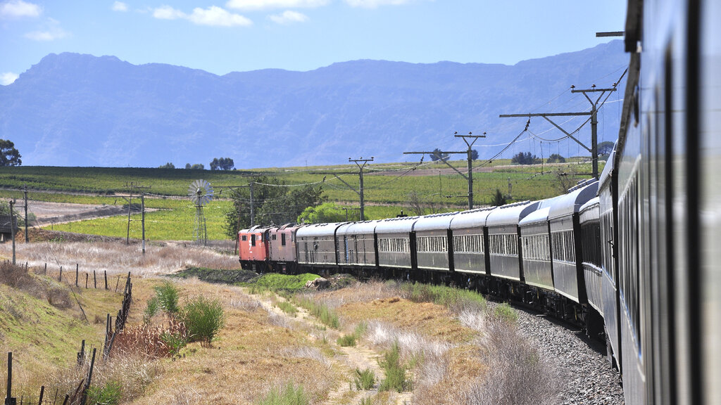 Ein langer Zug schlängelt sich durch eine fruchtbare Ebene, im Hintergrund eine schroffe Bergkette.