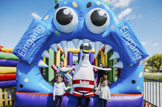 Ein Blaues Tor mit Kulleraugen ist der Eingang zur größten Hüpfburg Österreichs. Davor steht das Maskottchen und zwei Kinder.