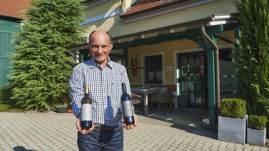 Winzer Manfred Edelmann präsentiert im Hof seines Betriebs zwei Weinflaschen, eine mit Chardonnay und eine mit einer Rotwein-Cuvée.