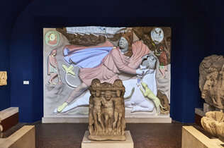 Figurengruppen aus Carnuntum und ein bemaltes Relief im Hintergrund.