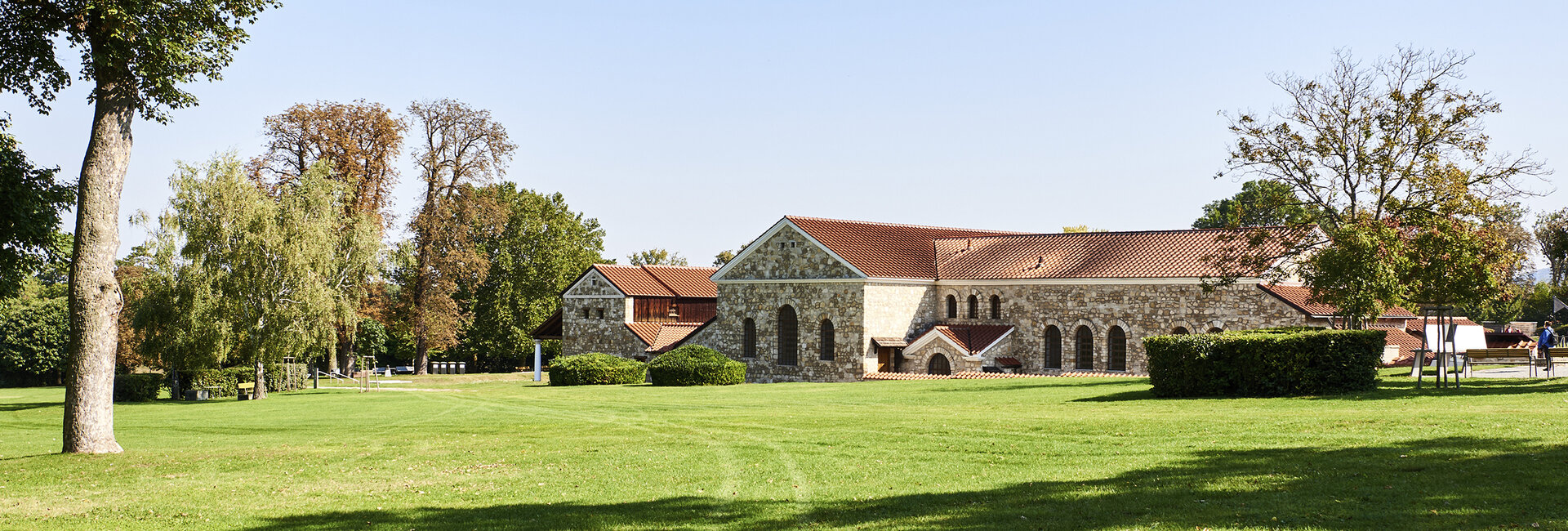 Hinter der grünen Wiese ist ein wieder aufgebautes großes römisches Haus aus Stein mit einem Ziegeldach zu sehen. 
