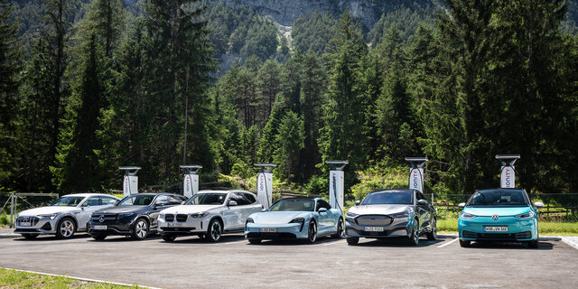 Sechs Elektroautos laden an sechs Schnellladern auf einem Parkplatz mit Bäumen im Hintergrund.