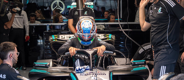 Ein Formel-1-Fahrer in voller Montur mit Rennanzug und -helm steht neben seinem Formel 1 Auto, um ihn herum arbeiten viele Teammitglieder.