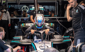 Ein Formel-1-Fahrer in voller Montur mit Rennanzug und -helm steht neben seinem Formel 1 Auto, um ihn herum arbeiten viele Teammitglieder.