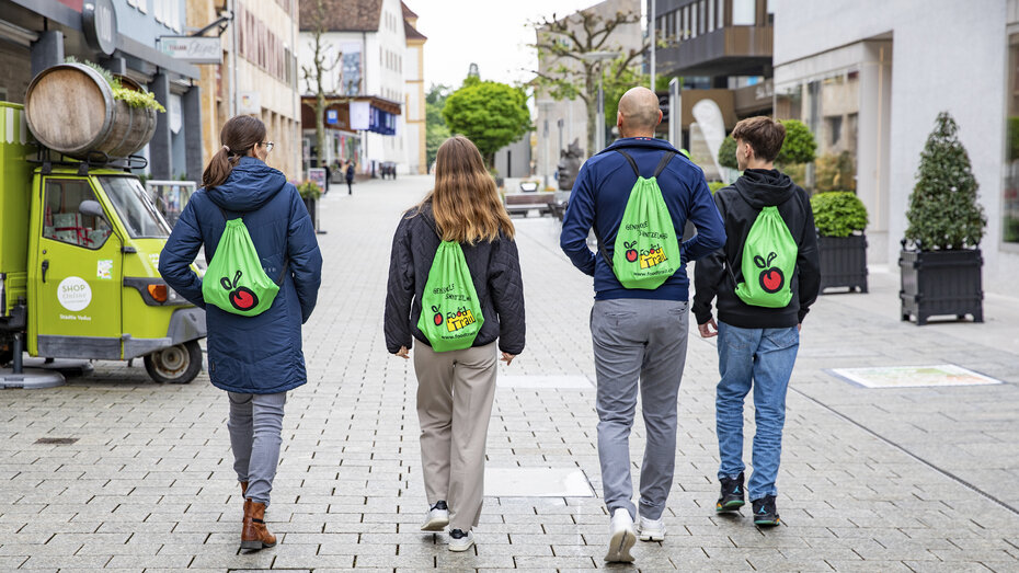 Vier Personen gehen in der Fußgängerzone und man sieht sie von hinten. Alle tragen den gleichen Rucksack.