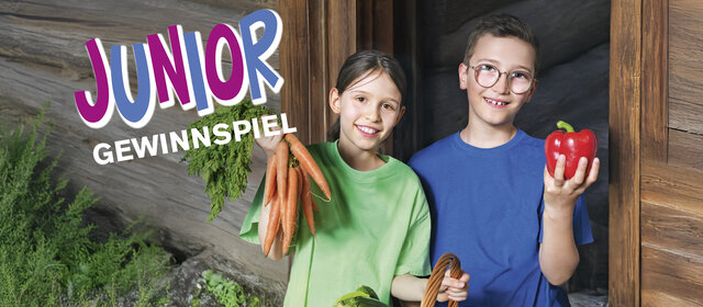2 Kinder (ein Mädchen und ein Bub) sitzen vor einer Blockhütte und halten Gemüse in den Händen.