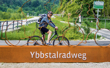Das Bild zeigt den auto touring-Redakteur, der auf dem Ybbstal-Fahrradweg unterwegs ist und vor einem großen orangefarbenen Sockel posiert, auf dem "Ybbstalradweg" steht. Auf dem Sockel befindet sich auch eine Metallskulptur von zwei Radfahrern. Im Hintergrund sieht man eine Straße in grüner Landschaft mit Bäumen und Hügeln. 