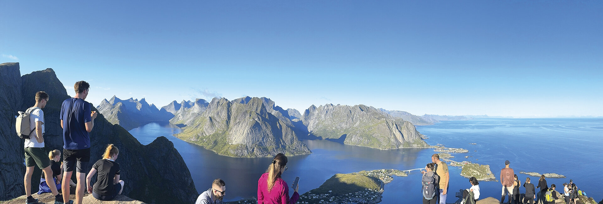 Spektakuläres Foto vom Berg: Blick auf die Fjorde, blauer Himmel und man sieht einige Wanderer die gerade hinunterschauen. 