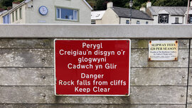 Ein Schild in walisischer Sprache, das vorm gefährlichen Wandern auf den Klippen warnt.