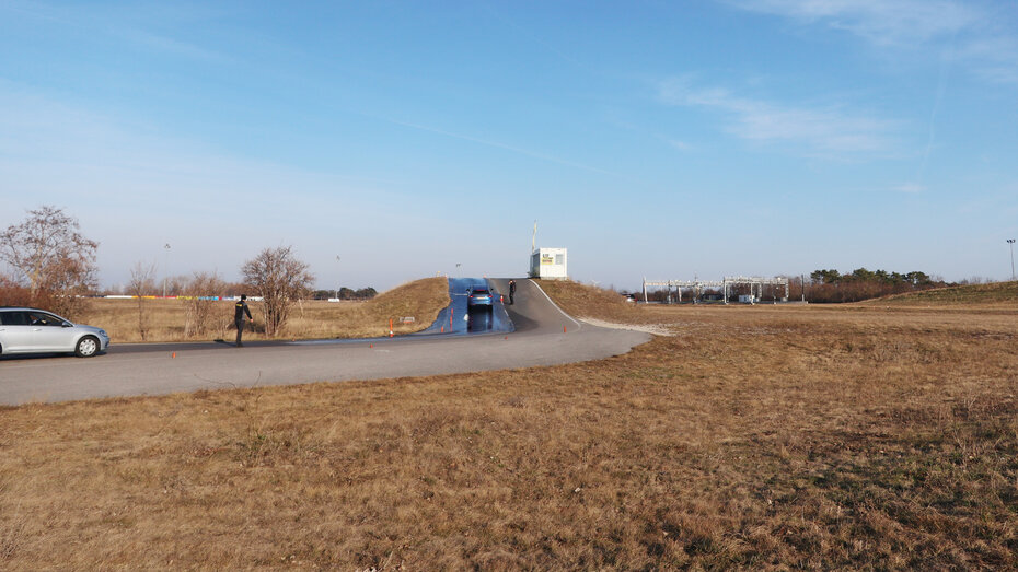 Eine große Grasfläche und eine nasse Straße, die über einen kleinen Hügel führt. Auf der Straße sind 2 Autos zu sehen. Trainingsübung  am Fahrtechnikgelände in Teesdorf.