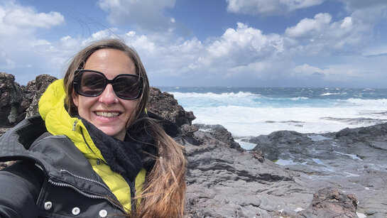 Eine Frau mit langen Haaren steht an der rauen Küste, im Hintergund das Meer.