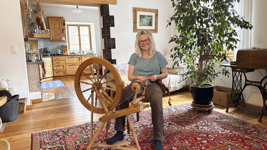 Eine Frau mit längeren grauen Haaren (Barbara Enk) sitzt vor einer alten Spindel und führt das Handwerk vor. Sie sitzt im Wohnzimmer, im Hintergrund ist ihre Küche zu sehen.