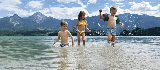 Drei kleine Kinder planschen im See, im Hintergrund die Bergkulisse.