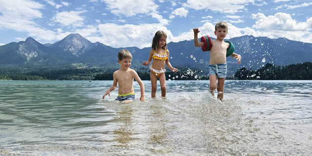Drei kleine Kinder planschen im See, im Hintergrund die Bergkulisse.