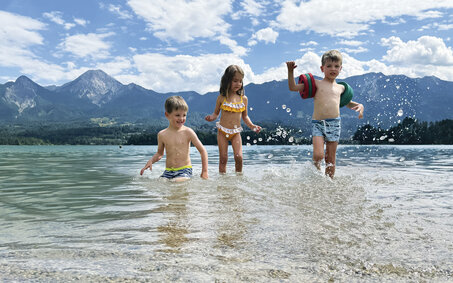 Drei kleine Kinder planschen im See, im Hintergrund die Bergkulisse.