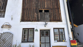 Weiße Hausmauer mit Eingangstür, daüber eine Holzmauer mit Guckloch, aus dem ein Alpaka rausschaut.