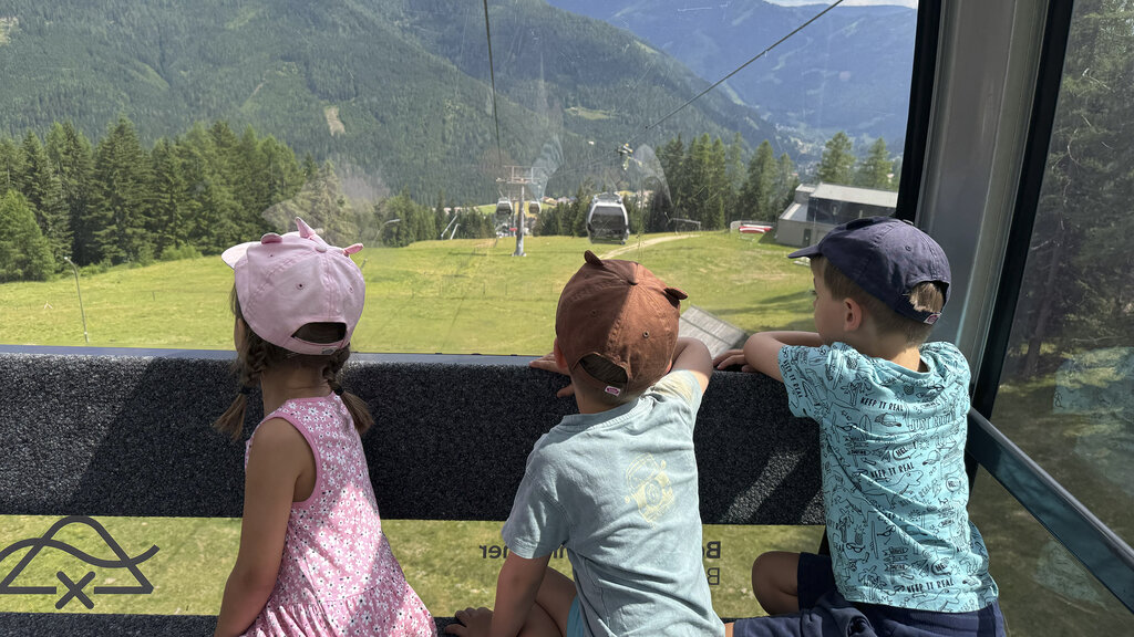 Drei Kinder sitzen in der Gondel und schauen auf die Almen hinunter.