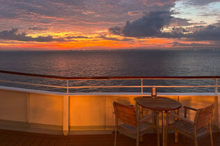 Zu sehen ist ein Tisch und zwei Stühle auf Deck an der Reling der MS Europa bei Sonnenuntgergang mit Blick auf das Meer.