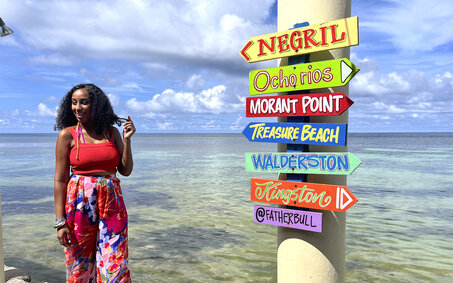 Bunte Ortsschilder am Meer und eine Dame steht daneben.