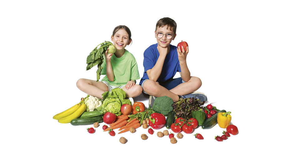 Ein Mädchen mit grünem T-Shirt hält einen Kohlrabi in der Hand, daneben sitzt ein Bub im blauen T-Shirt und hält einen roten Apfel. Vor ihnen liegt Obst und Gemüse.