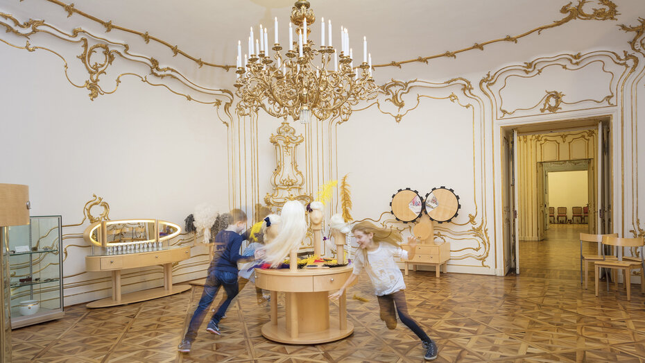Kinder laufen im Schloss Schönbrunn in einem barocken Raum um einen Tisch herum.