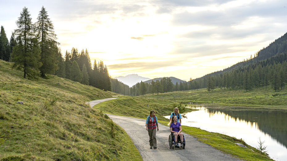 Zwei Frauen und eine Frau im Rollstuhl spazieren entlang eines Weges neben einem See, im Hintergrund ein Hügel.