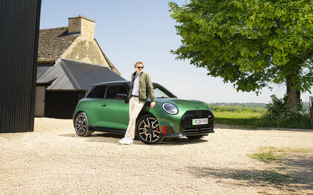 Ein grüner Mini steht auf einem Schotterparkplatz, davor lehnt eine Person.