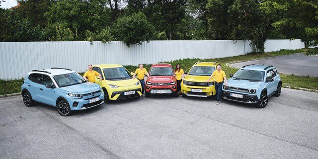 Fünf kleine Elektroautos nebeneinander aufgereiht, stehend von vorne, mit vier Personen