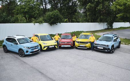 Fünf kleine Elektroautos nebeneinander aufgereiht, stehend von vorne, mit vier Personen