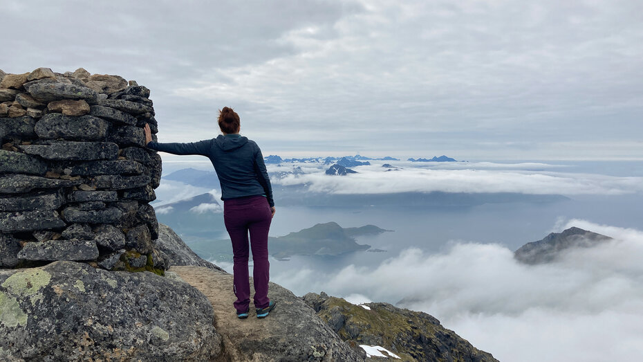 Rückansicht einer Frau, die am Gipfel steht und auf ein wolkenverhangenes Panorama blickt.