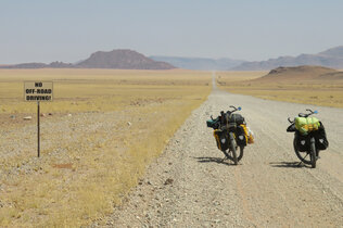 Zwei mit Reisegepäck beladenen Fahrräder stehen auf einer Schotterstraße in Namibia vor der weiten Landschaft, im Hintergrund felsige Hügel.