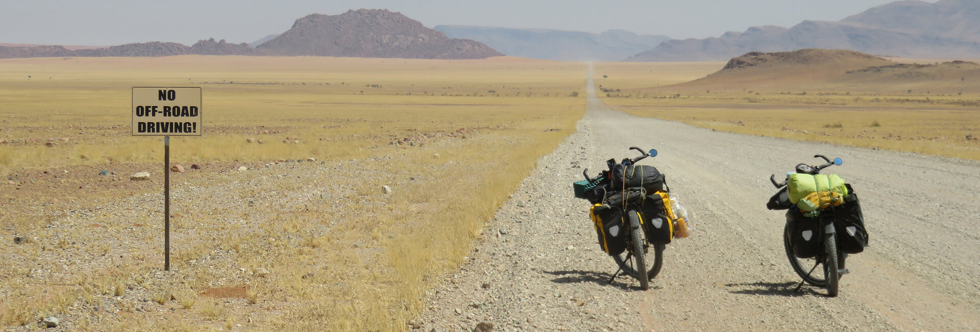 Zwei mit Reisegepäck beladenen Fahrräder stehen auf einer Schotterstraße in Namibia vor der weiten Landschaft, im Hintergrund felsige Hügel.