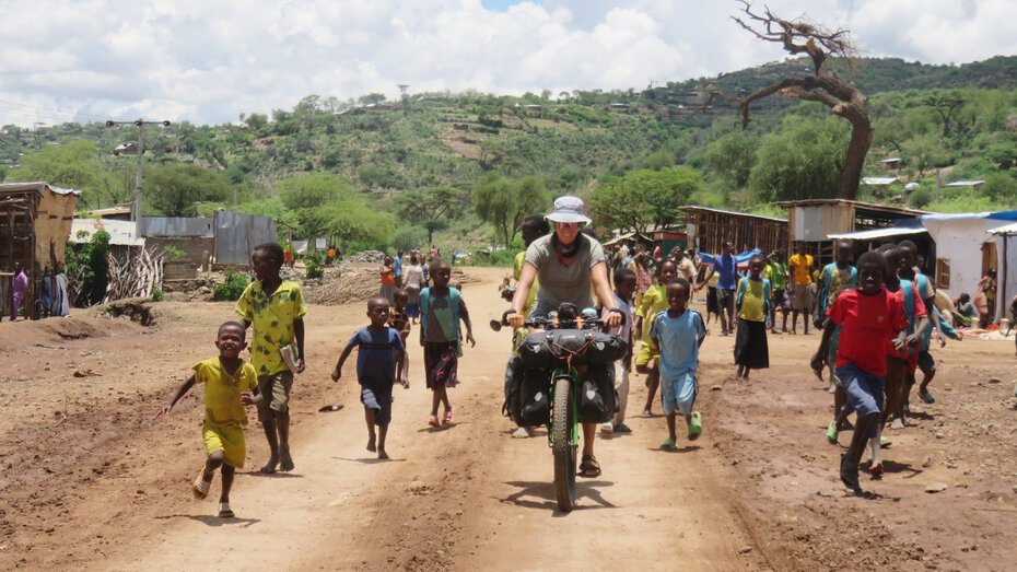 Eine Radreisende fährt am Rad auf einer sandigen Straße durch ein Dorf in Äthiopien. Rund um sie sind Einheimische zu sehen.