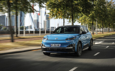 Ein blauer Nissan Micra 2025 fährt auf einer Straße, hinter der sich Bäumen und moderne Gebäude befinden.