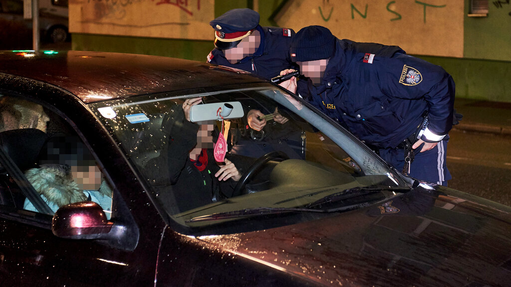 Eine Fahrzeugkontrolle durch zwei Polizisten bei Nacht, sie leuchten mit einer Taschenlampe und kontrollieren den Ausweis des Fahrers. Im Auto sitzen zwei Personen.